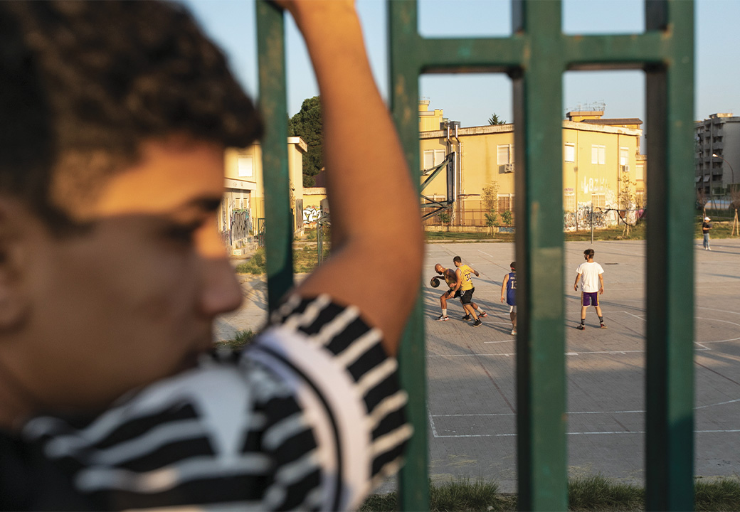 Un ragazzo guarda un campetto da basket attraverso delle sbarre